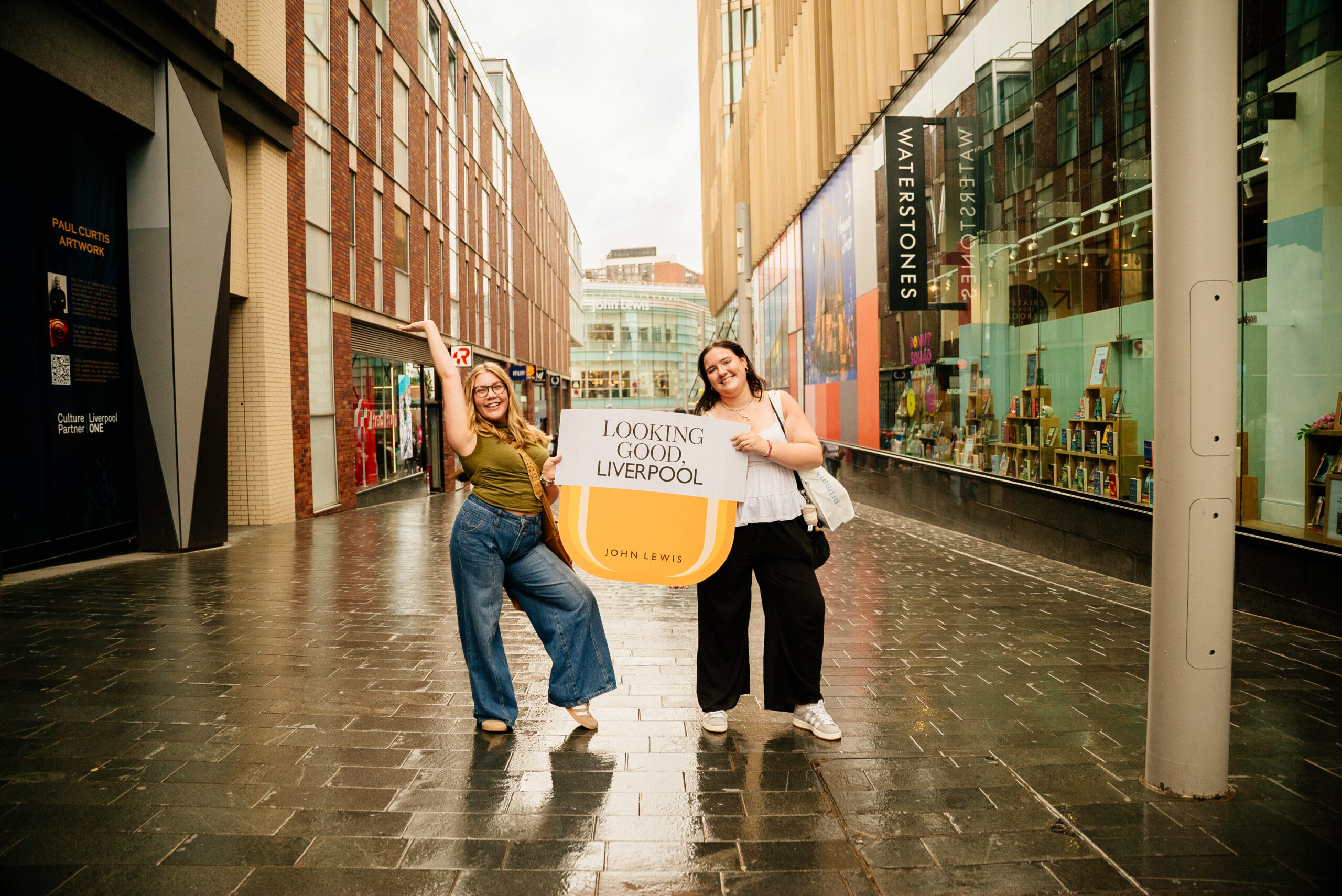 Two smiling women stand on a wet street in Liverpool, holding a large sign that reads Looking Good Liverpool (John Lewis) in a shopping area with shops and glass windows behind them.