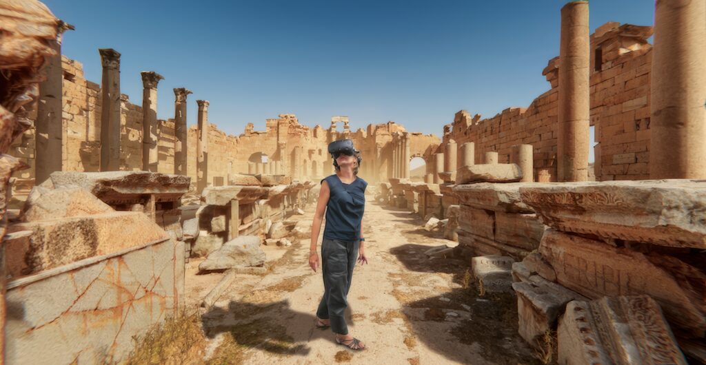 A person wearing casual clothes and a hat walks through ancient stone ruins with tall columns under a bright blue sky, surrounded by broken pillars—a scene reminiscent of immersive marketing bringing history to life.