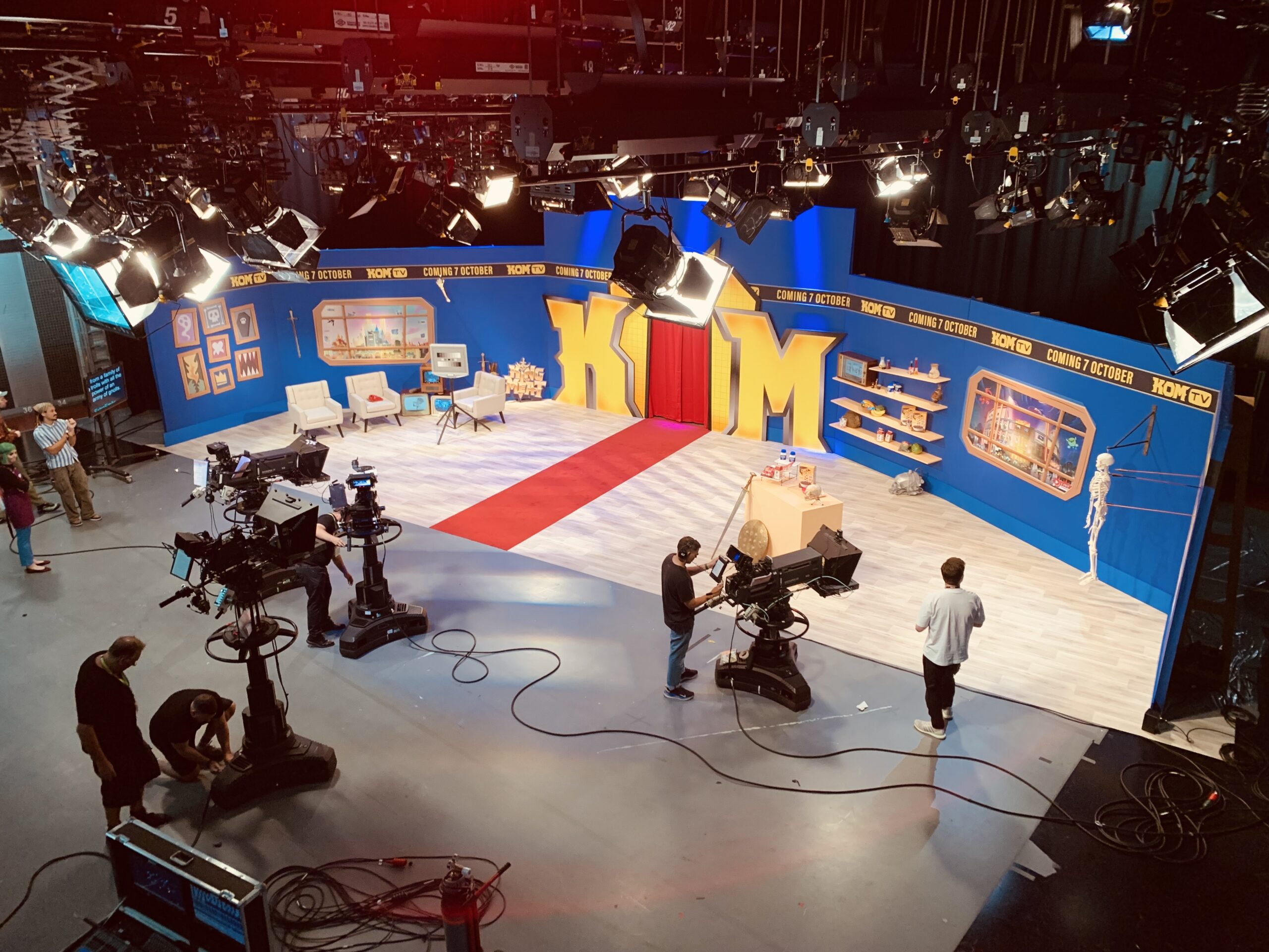 A brightly lit TV studio set with cameras, crew, and a colorful backdrop featuring the large yellow letters KUM. There’s a red carpet, chairs, props, and shelves, creating a lively, playful environment.