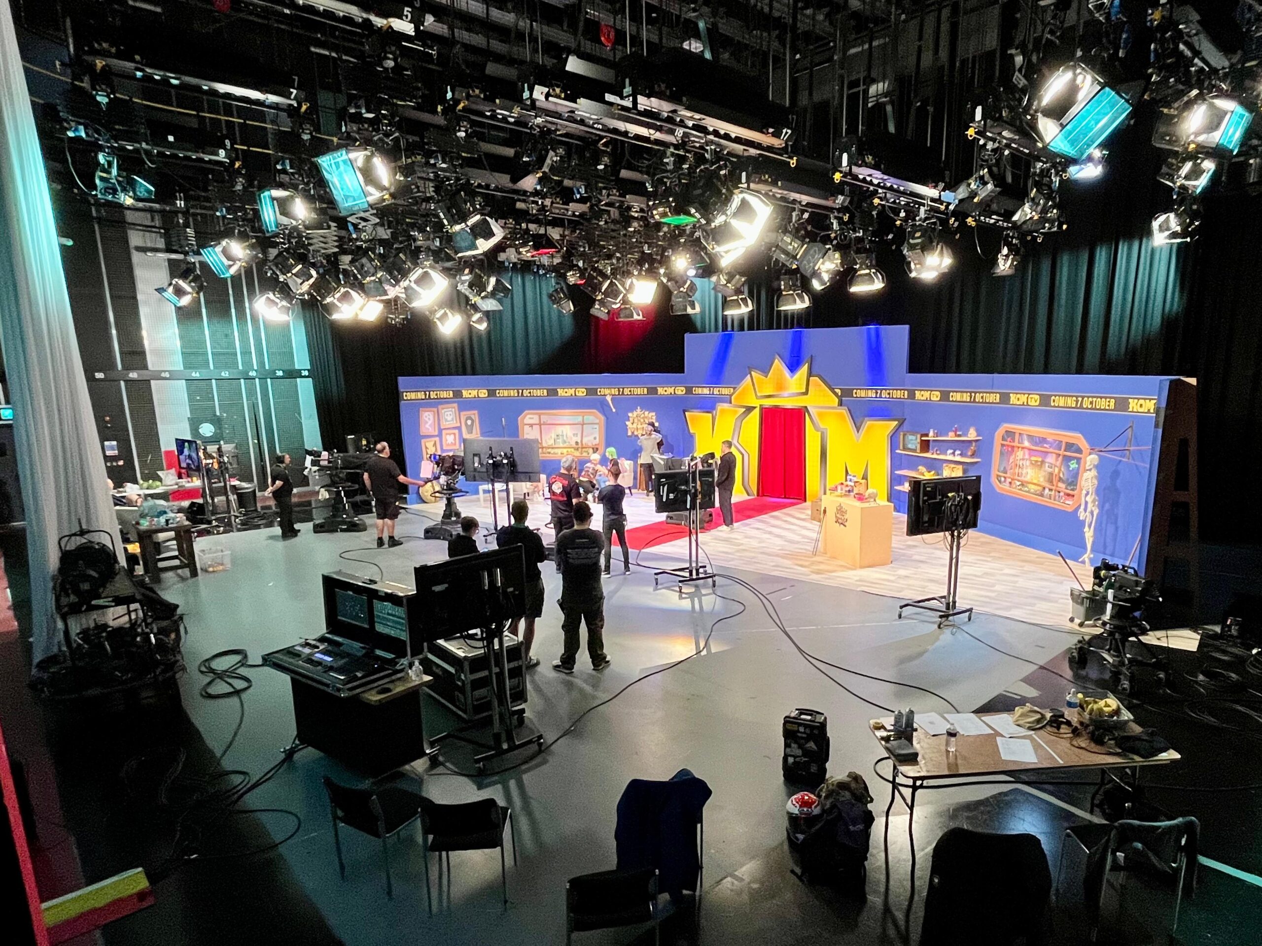 A brightly lit TV studio set with cameras, crew members, and equipment. The set features a blue and gold castle-like backdrop with a crown design, stage props, and workspaces, with technicians and staff preparing for filming.