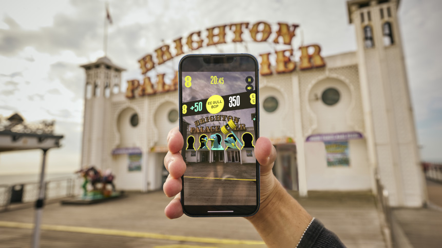 A hand holds a smartphone in front of the Brighton Palace Pier entrance, displaying an augmented reality game overlaying digital graphics onto the real scene.