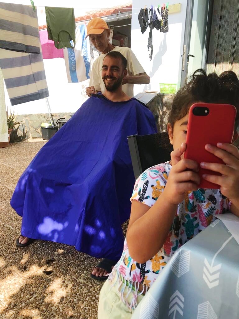 A man sits outdoors with a blue cape, smiling as an older man cuts his hair. In the foreground, a child in a colorful shirt holds a red smartphone, appearing to take a photo. Towels and laundry hang in the background.