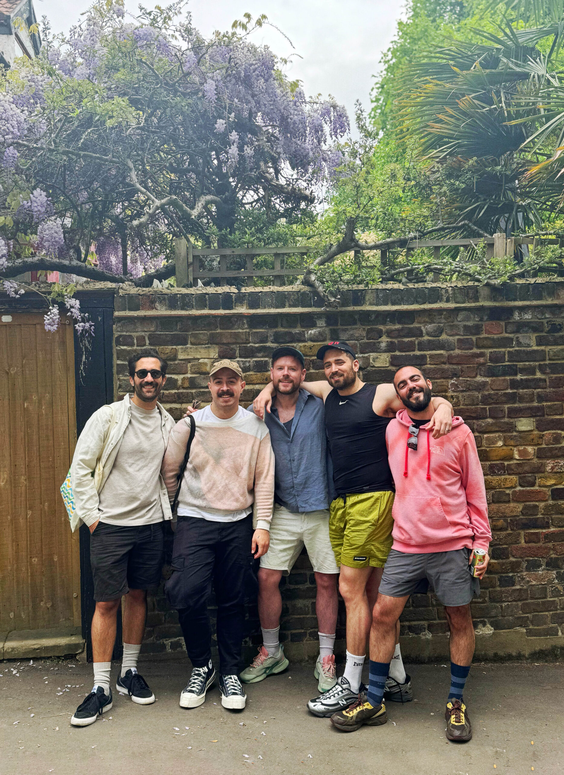 Five men stand close together, smiling in front of a brick wall with purple wisteria flowers overhead. They are dressed casually in shorts and light jackets, appearing relaxed and happy outdoors.