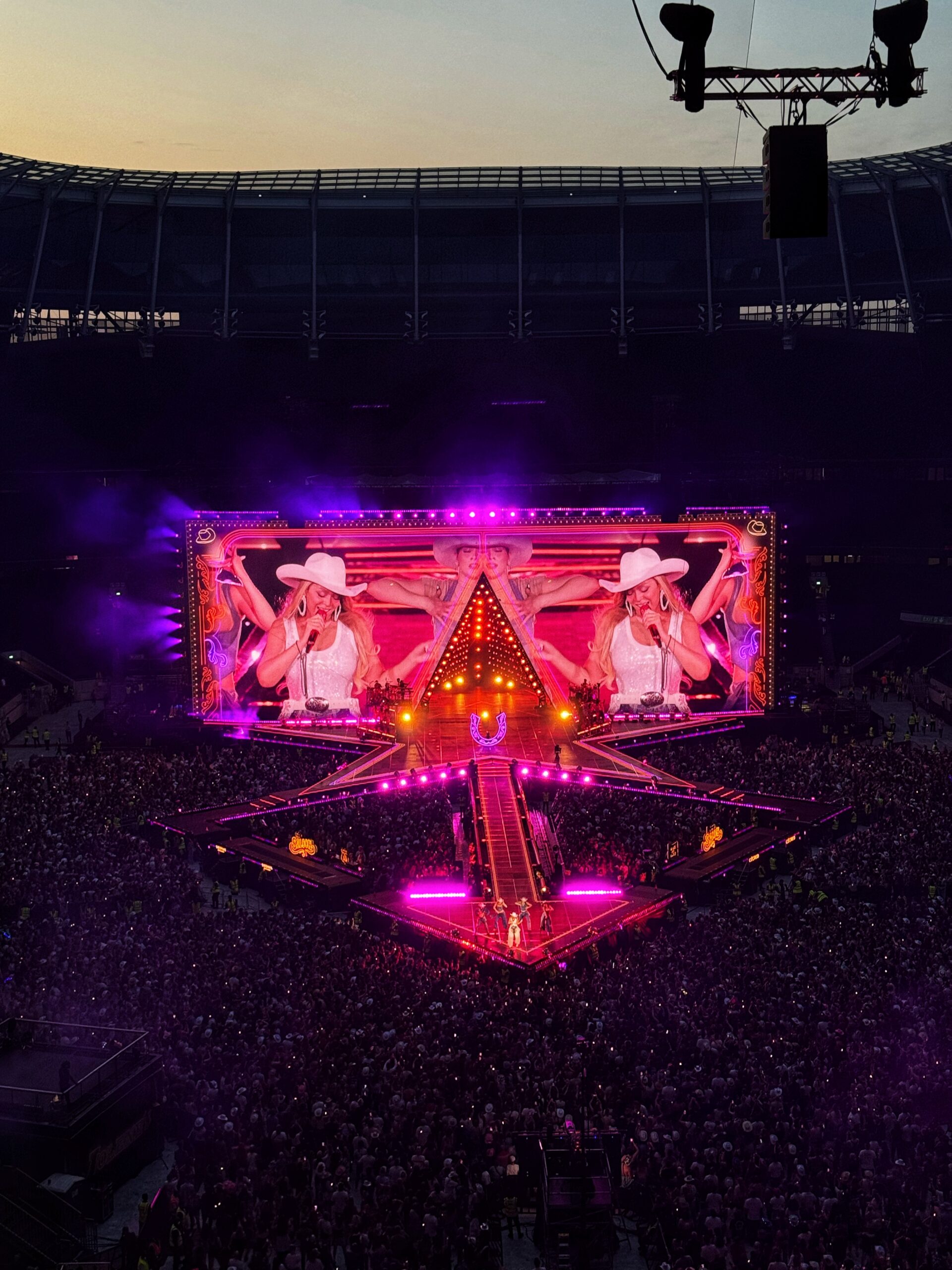 A large crowd watches a performer on a brightly lit stage with pink lights and a big screen showing the singer, who wears a hat, at a concerts in a stadium at sunset.
