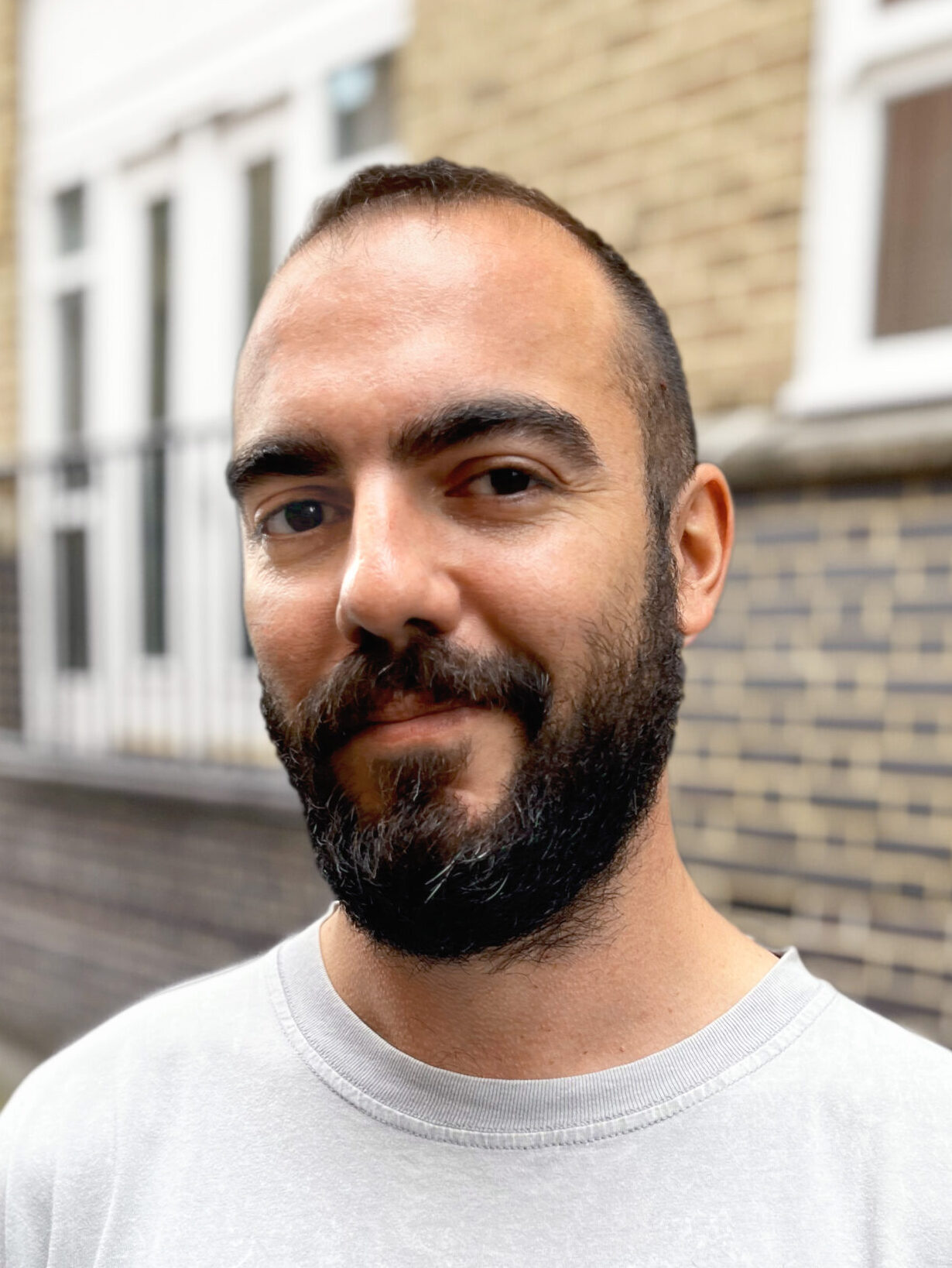 A man with a beard and short hair stands outside in front of a brick building, wearing a light gray T-shirt that says PARADISE CHASER. He is looking at the camera and smiling slightly.