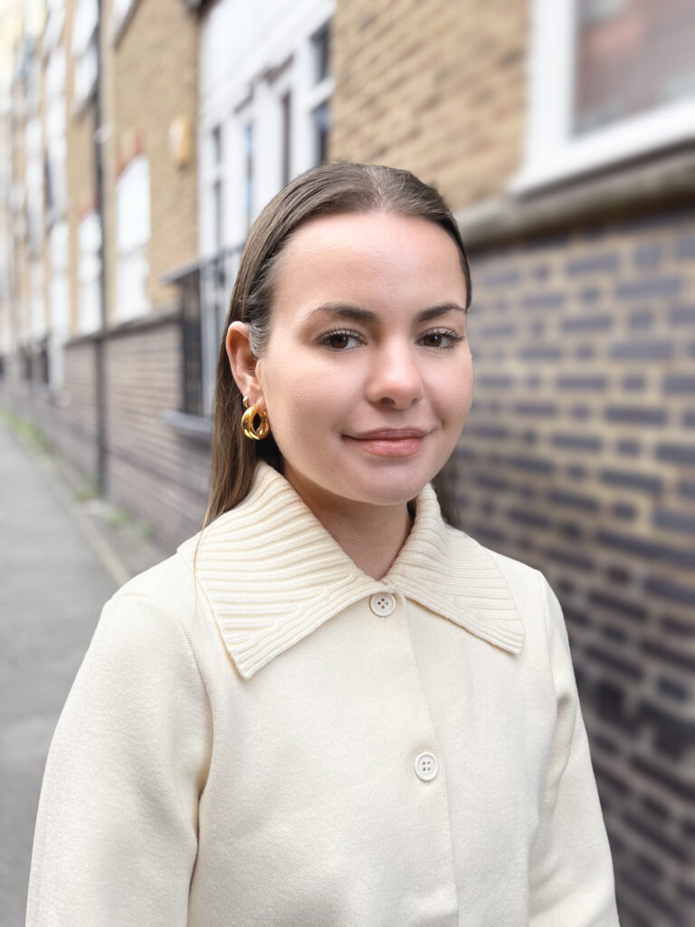 A woman with straight brown hair, wearing gold hoop earrings and a cream-colored coat with a large collar, stands on a city sidewalk beside a brick building. She is smiling slightly and looking at the camera.