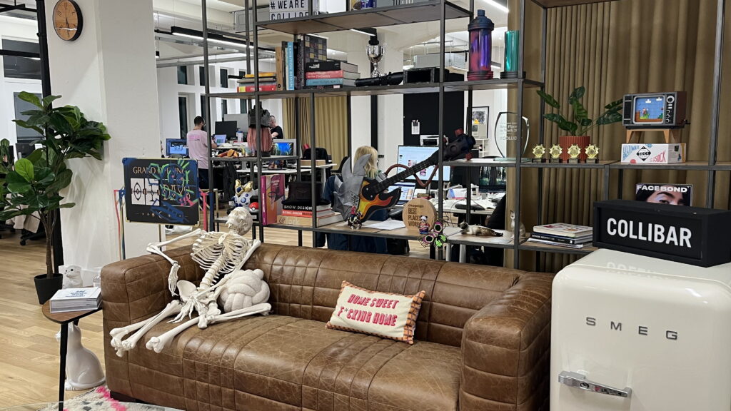 A skeleton sits on a brown leather couch with a Home Sweet Second Home pillow in a modern office filled with desks, plants, colorful decor, and a white SMEG fridge. People work at computers in the background.