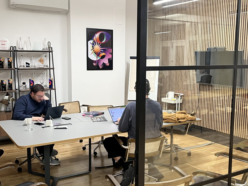Two people are working on laptops at a conference table in a modern office with glass walls. Shelves with awards and art are visible, along with a coat on a chair and a whiteboard in the background.