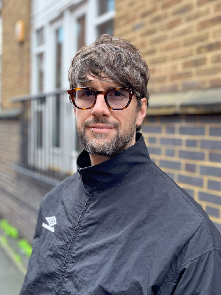 A man with short, wavy brown hair and a beard wears sunglasses and a black jacket with the Umbro logo, standing in front of a brick wall outdoors.