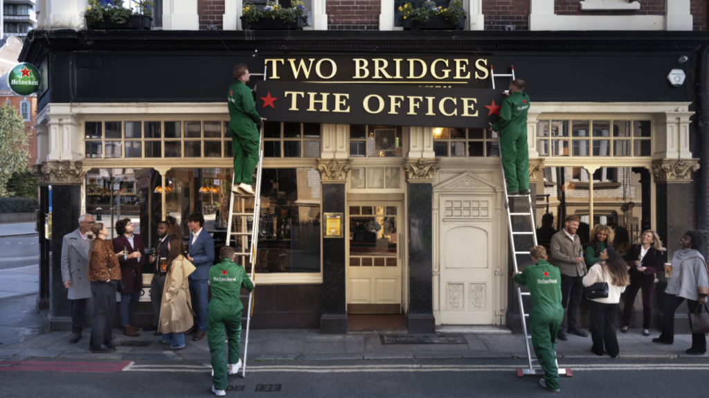 Four workers in green uniforms on ladders, orchestrated by a brand experience agency, change a pub sign from TWO BRIDGES to THE OFFICE as pedestrians watch and gather outside. The street scene feels busy and social.