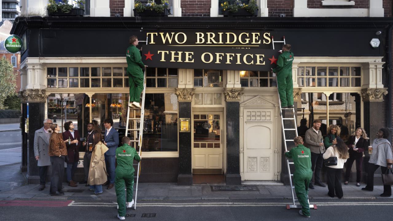 Four workers in green uniforms on ladders, orchestrated by a brand experience agency, change a pub sign from TWO BRIDGES to THE OFFICE as pedestrians watch and gather outside. The street scene feels busy and social.