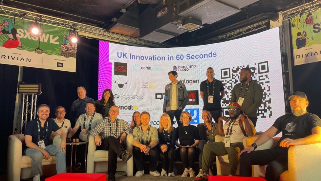 A group of people pose on a stage in front of a screen displaying UK Innovation in 60 Seconds with logos, a QR code, and event branding, highlighting discussions around AI in marketing. Some are seated on couches, others stand behind them, all smiling at the camera.