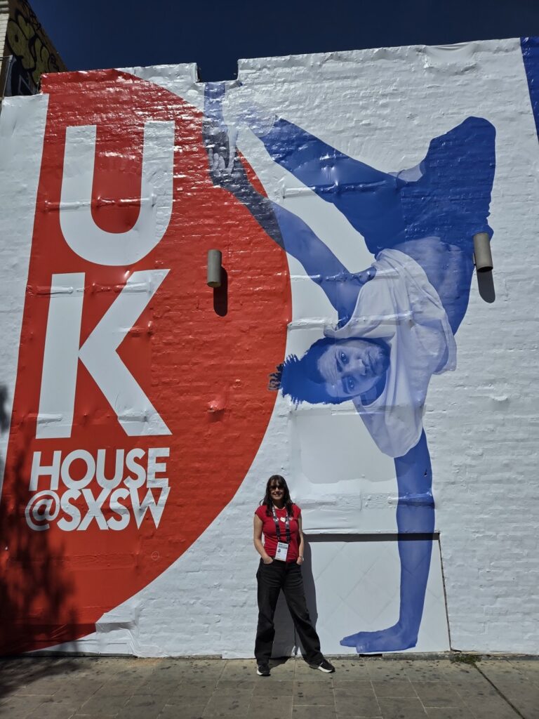 A person stands before a large mural reading UK House @ SXSW, featuring a blue-toned image of a breakdancer performing a handstand. The mural, painted on a white brick wall, hints at themes like AI in marketing and creative innovation.