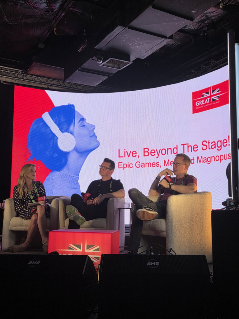 Three people sit on stage chairs with microphones in hand, speaking at an event about AI in marketing. Behind them, a large screen displays a woman with headphones and the text: “Live, Beyond The Stage! Epic Games, Meptik and Magnopus.”.