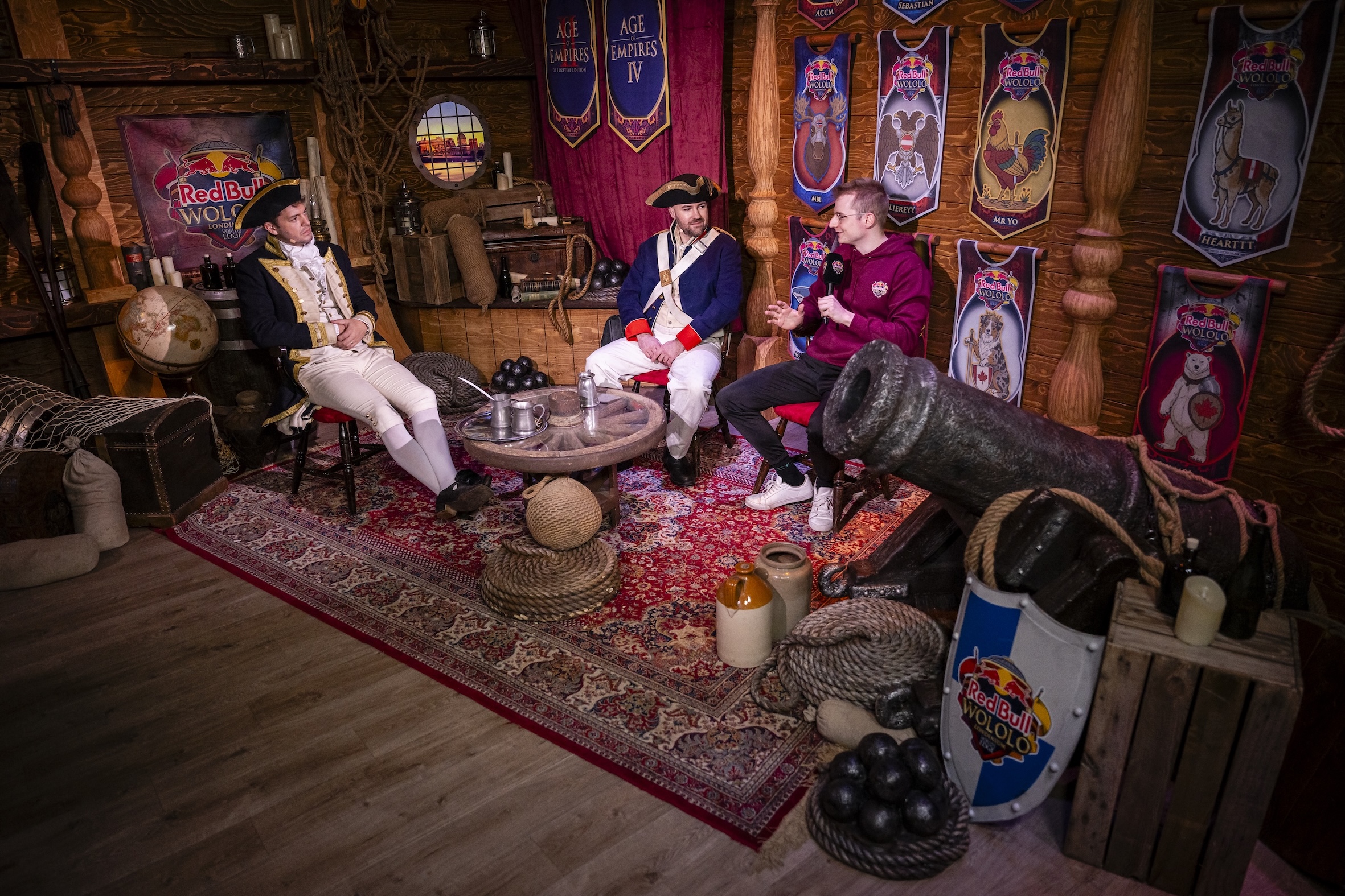 Three men in historical and modern costumes sit on chairs in a pirate-themed room adorned with Age of Empires IV banners, a cannon, barrels, and Red Bull branding. The cozy wooden tavern sets the stage for the exciting Red Bull LOIO event.