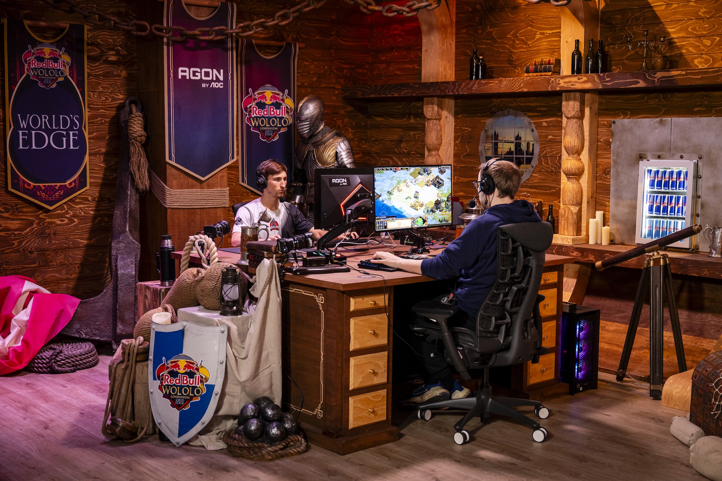 Two people compete in an esports tournament in a medieval-themed room, each at their own computer desk with gaming gear. Red Bull LOIO banners and props set the scene, while a fridge stocked with drinks adds to the excitement.