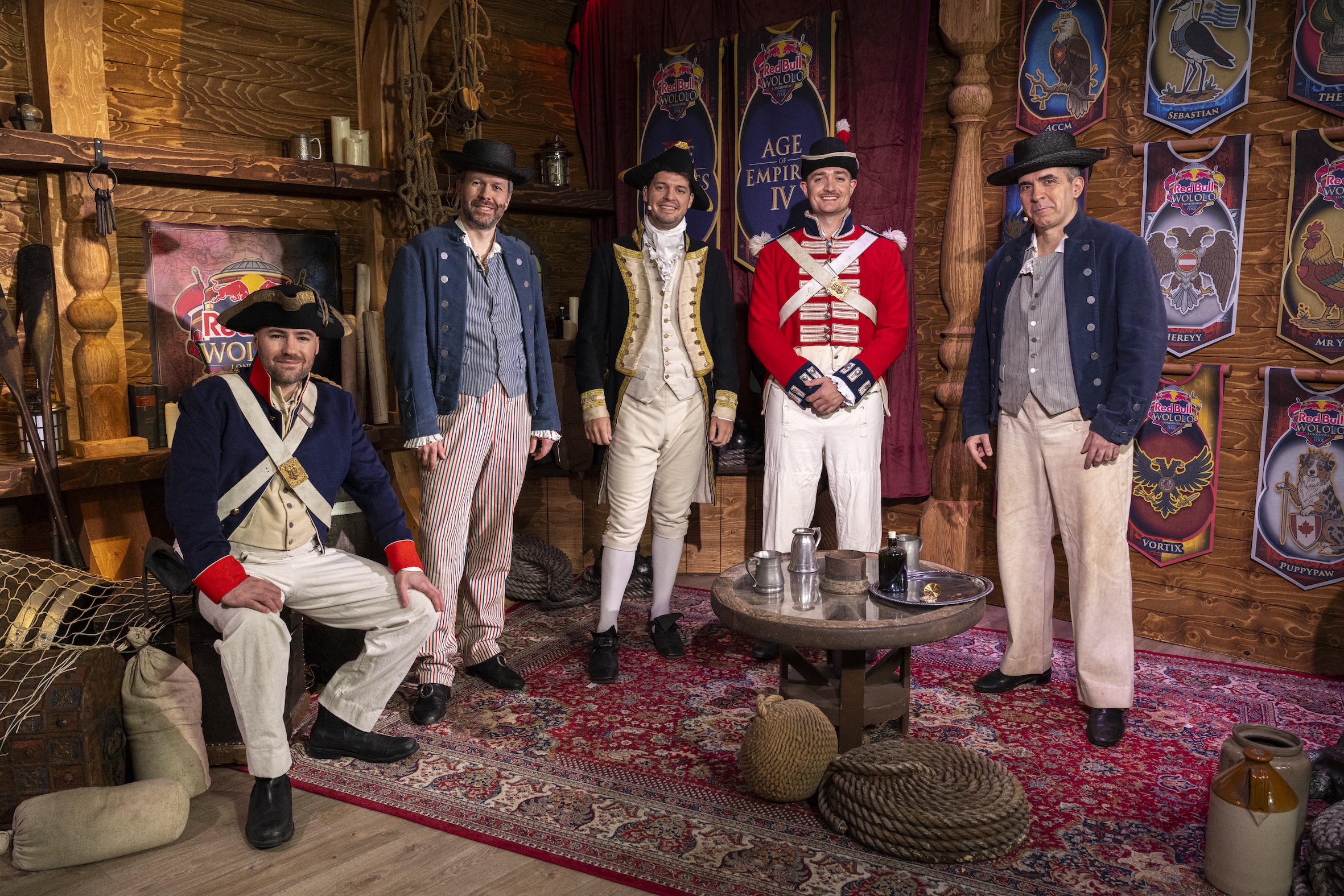 Five men wearing historical military uniforms from the 18th century pose inside a wooden room decorated with banners, shields, and Red Bull LOIO props, suggesting a lively reenactment or themed event.
