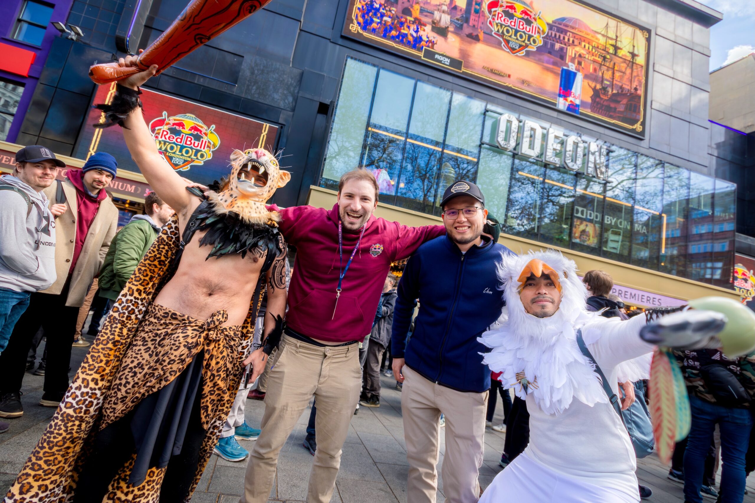 Two people in animal costumes and two casually dressed men pose and smile together outside a cinema with bright Red Bull LOIO and VOLOLO advertisements in the background. A crowd gathers behind them.