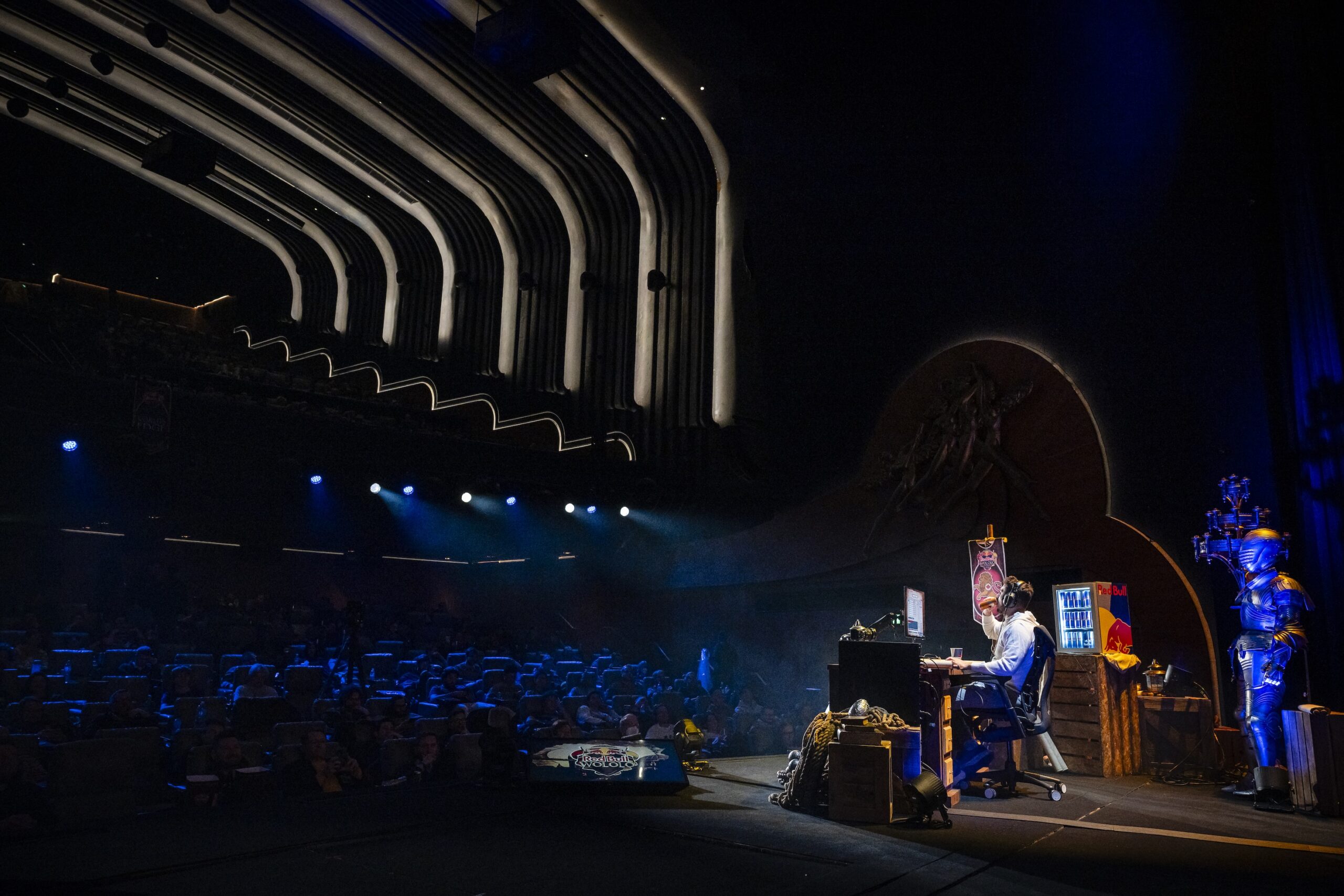 A person sits at a desk on a theater stage, illuminated by spotlights, with Red Bull LOIO signage in the decorated background and knight armor beside them. The audience is visible in the darkened seats of the modern auditorium.