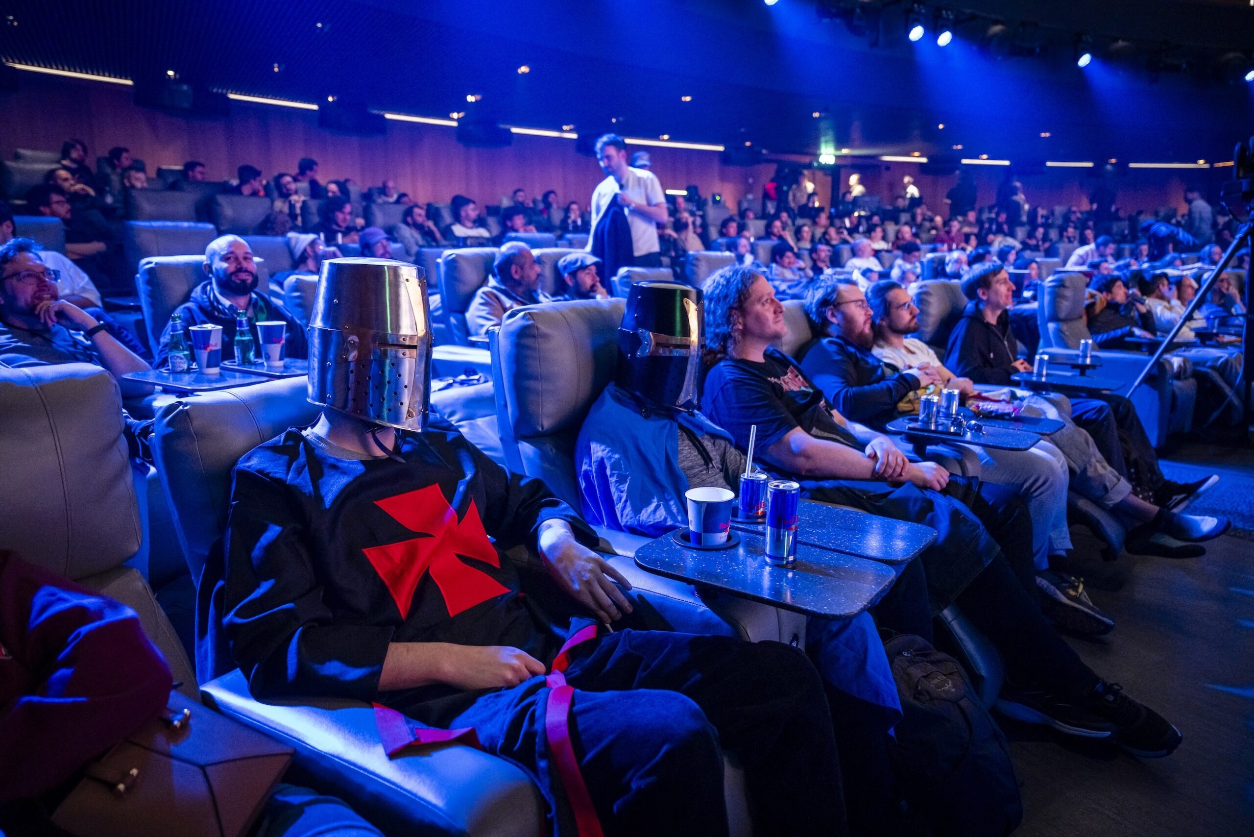 Audience members sit in a theater, with two people in the front row wearing medieval knight costumes and metal bucket helmets. The lively crowd, illuminated by blue lighting, buzzes with anticipation for the Red Bull LOIO event.