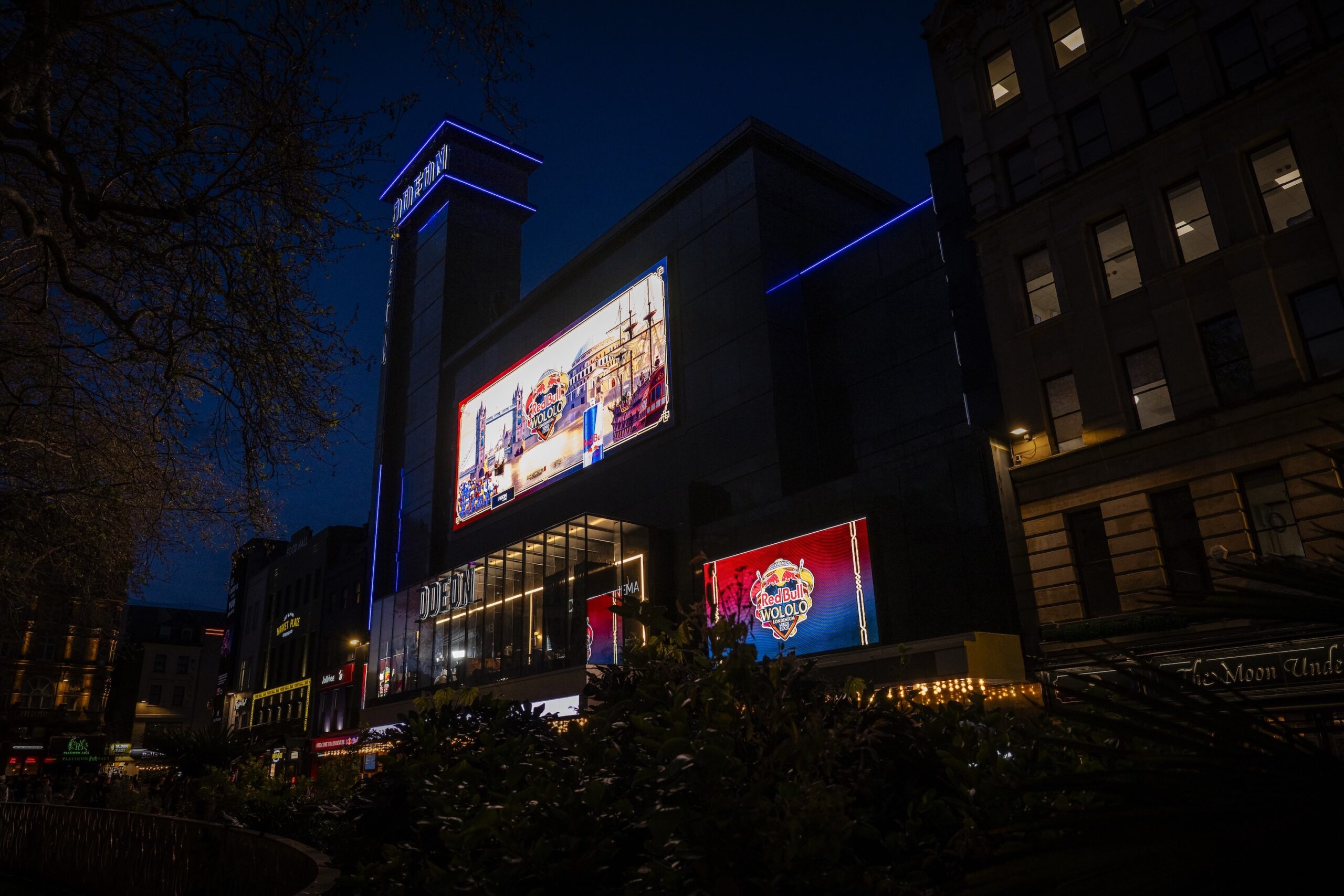 A city building at night is illuminated by colorful neon lights and two large, brightly lit digital billboards displaying vibrant Red Bull LOIO advertisements. Trees and other buildings surround the scene.
