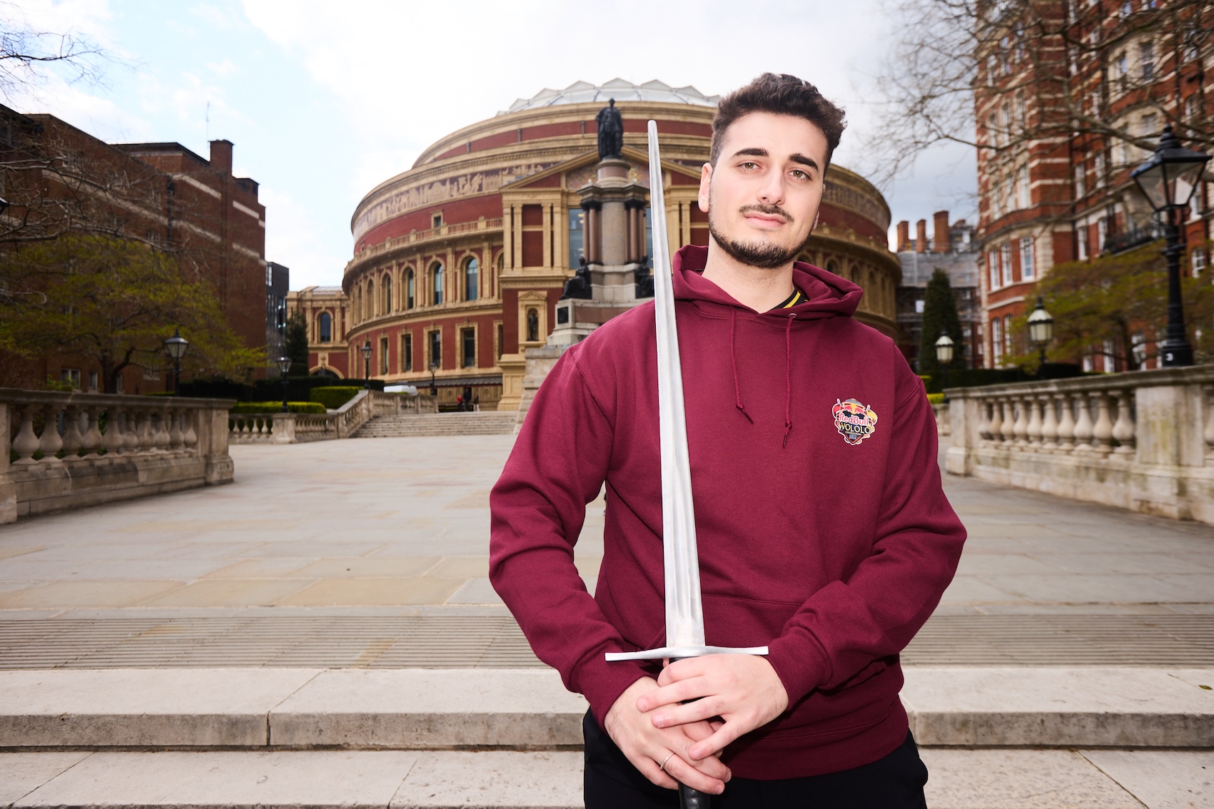 A young man in a maroon hoodie, possibly gearing up for Red Bull LOIO, holds a sword upright in both hands, standing in front of the Royal Albert Hall in London on a cloudy day.