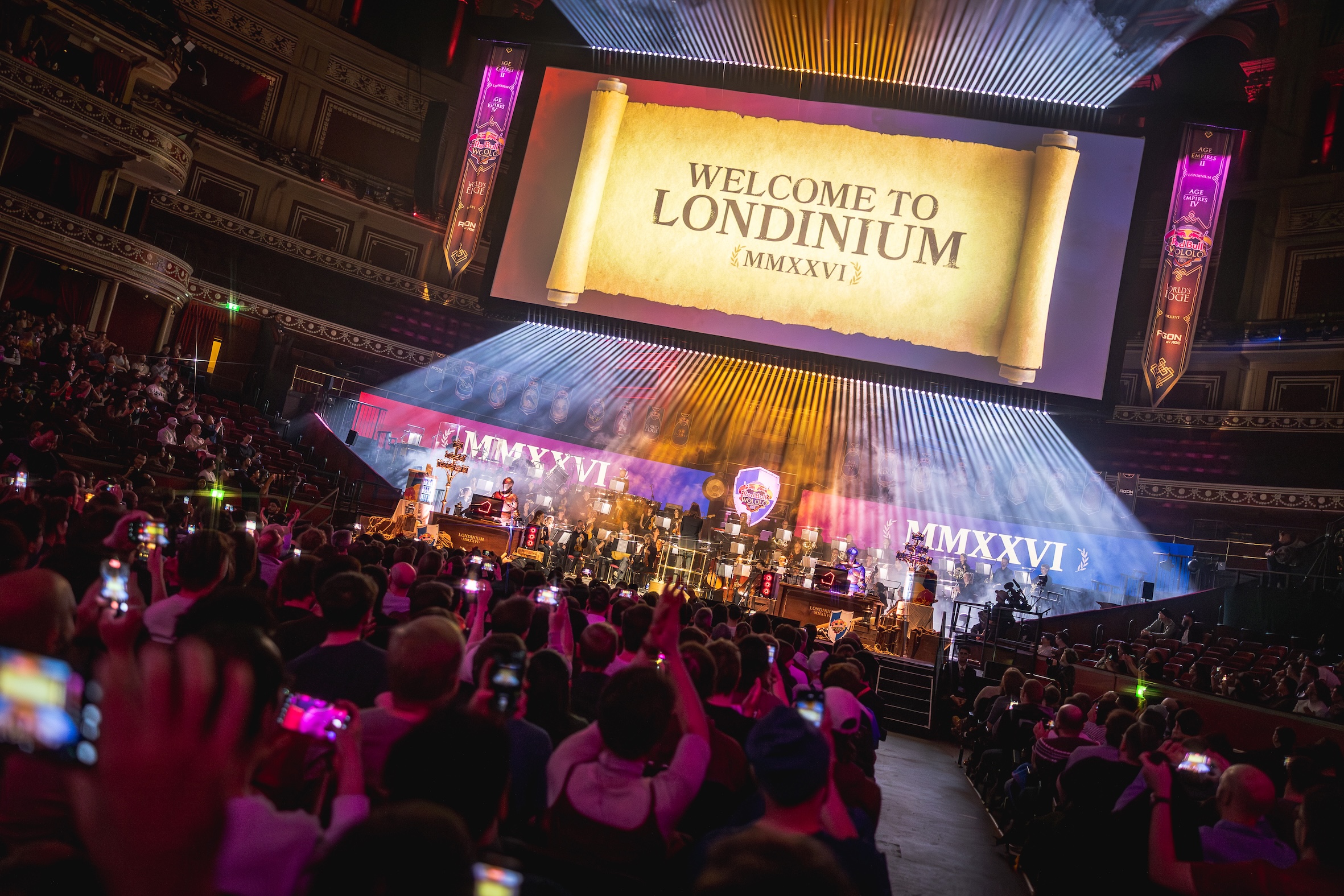 A large crowd watches a stage show with colorful lights and performers. A giant screen displays WELCOME TO LONDINIUM {MMXXVI} above the stage as the Red Bull LOIO event energizes the grand indoor venue, with the audience capturing every moment.