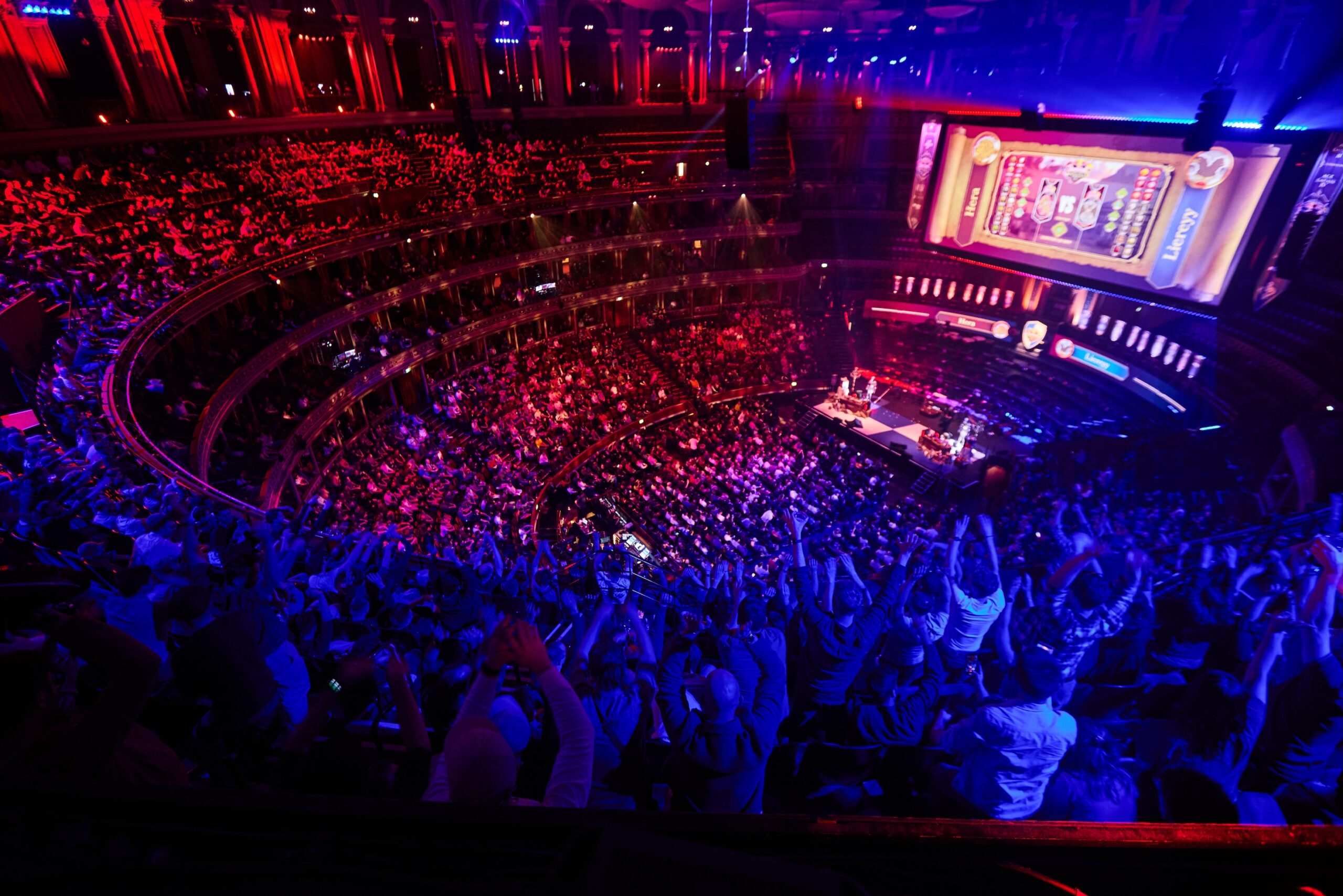 A large, vibrant crowd fills an arena with multiple balconies, cheering and illuminated by colorful lights. A giant screen displays a video game as players compete on stage below at the electrifying Red Bull LOIO event.