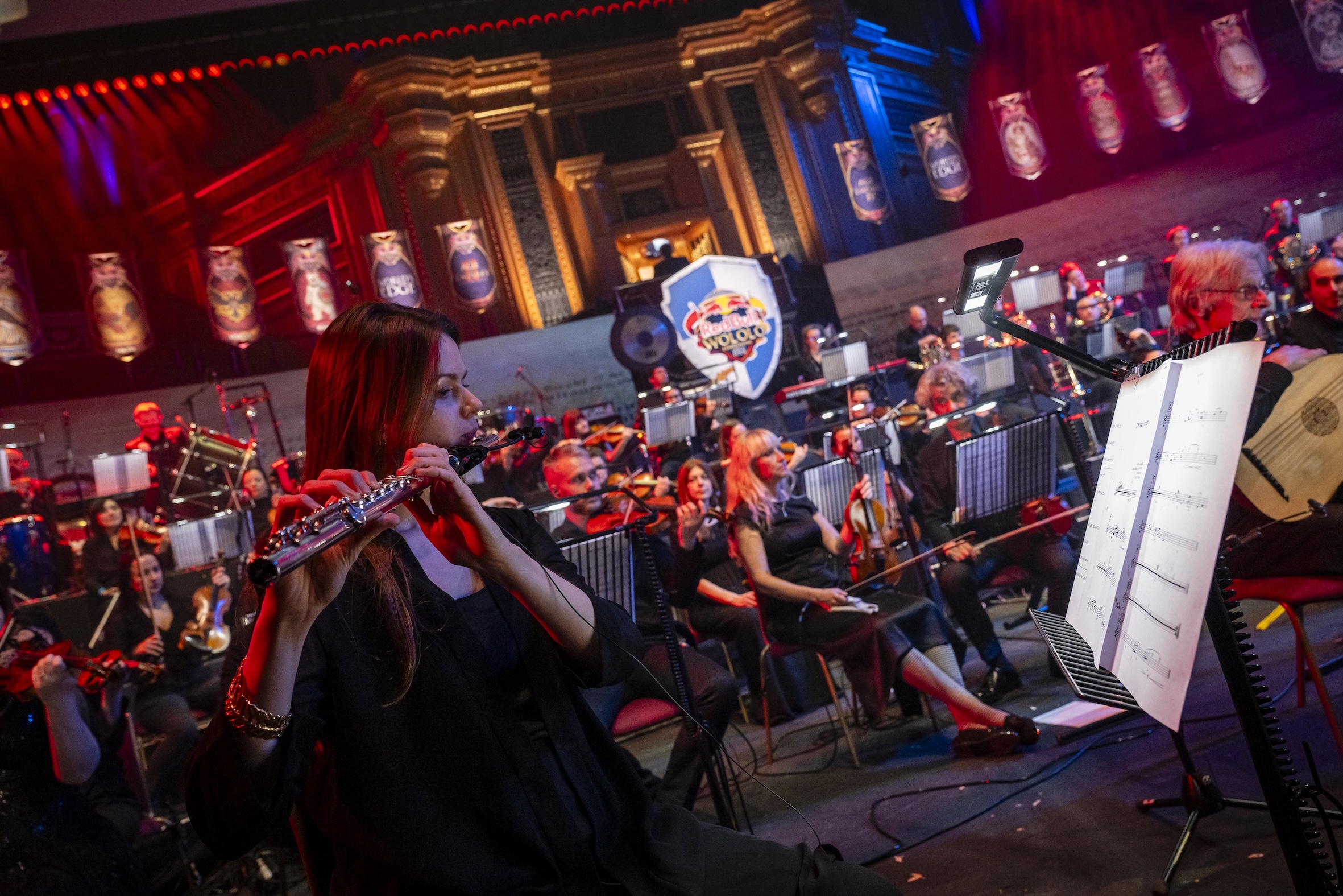 A female flutist plays in the foreground of a large orchestra on stage at Red Bull LOIO, with dramatic red and blue lighting and ornate golden architecture in the background. Sheet music and other musicians are visible.