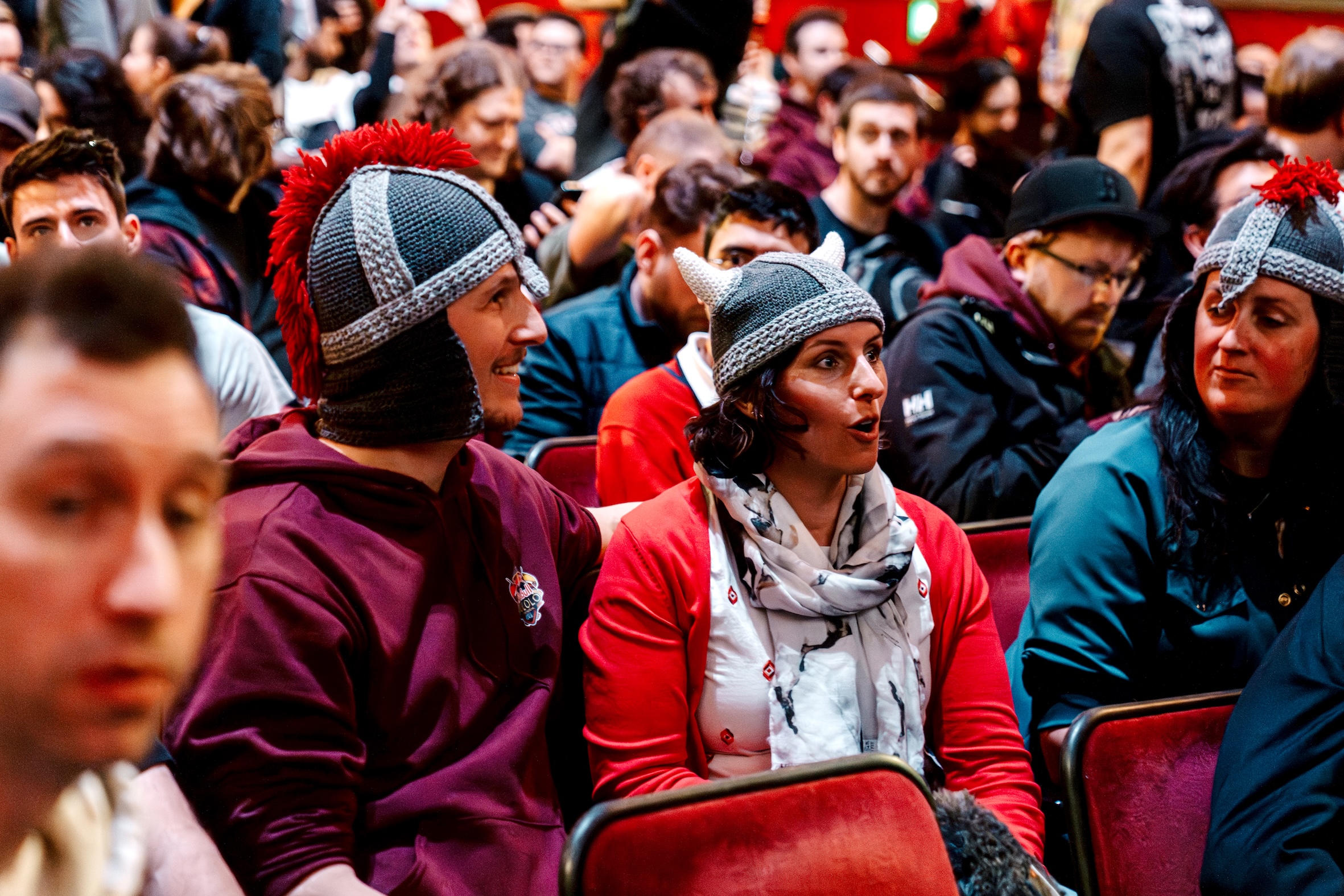 A crowd sits in red chairs at an indoor Red Bull LOIO event. In the foreground, two people dressed in knitted knight helmets and maroon clothing are engaged in animated conversation, while others watch or chat in the background.