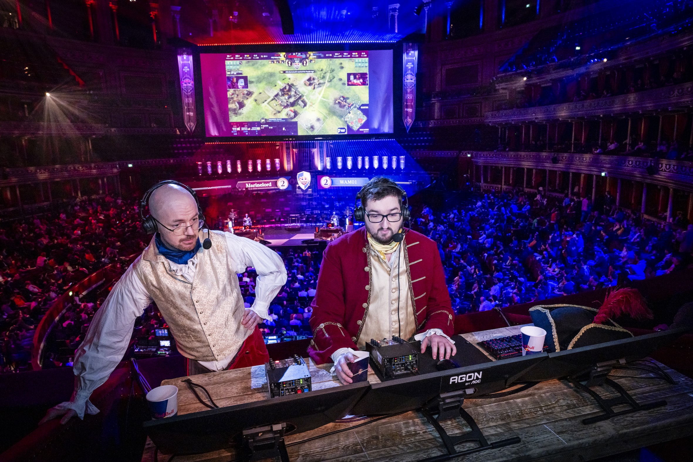 Two men in historical costumes work at computers on stage during Red Bull LOIO, with a large screen displaying a game behind them. The auditorium is filled with a crowd, lit by vibrant red and blue lights.