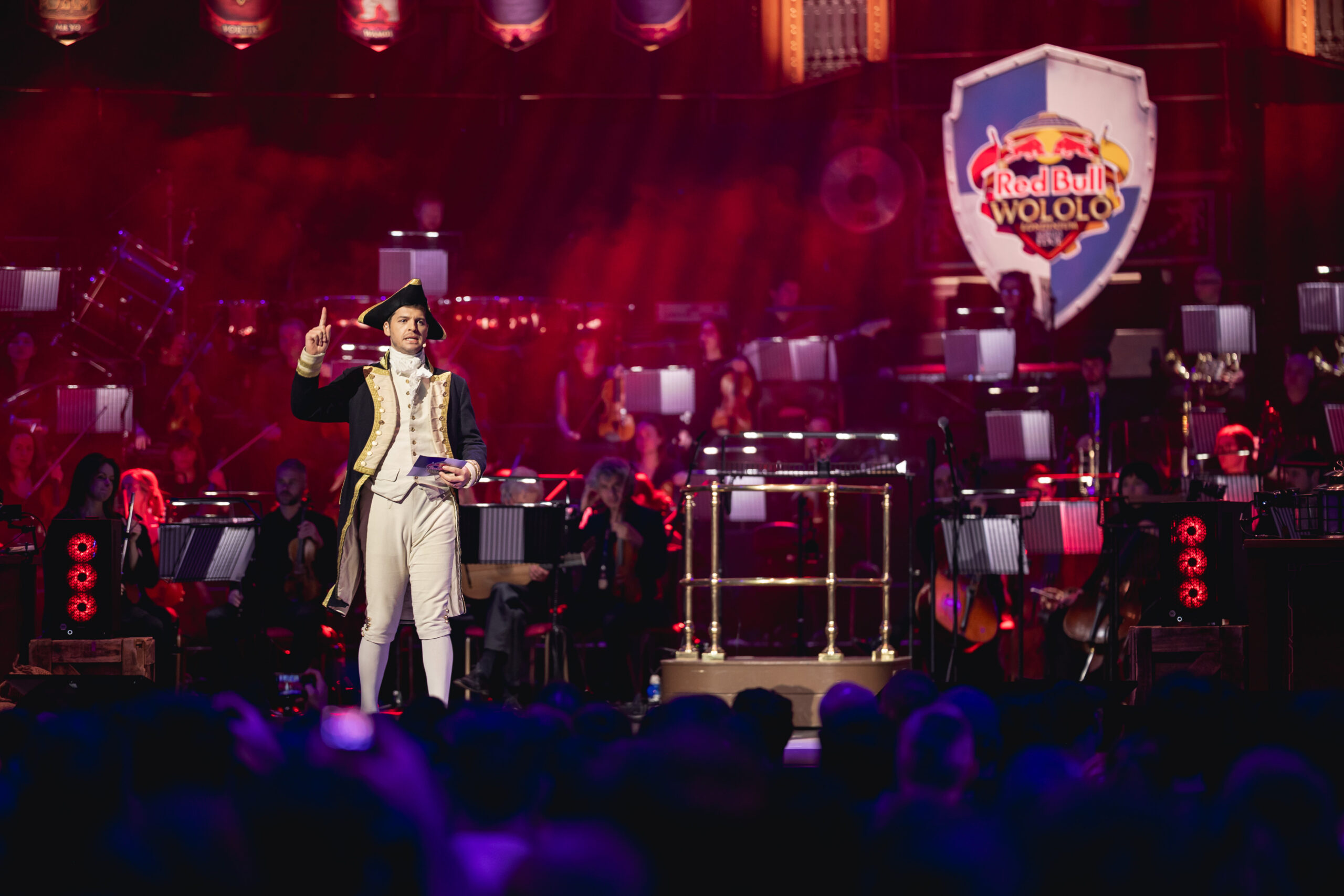 A person in historical costume stands on stage, gesturing, in front of an orchestra. The stage is lit with red and purple lights, and a large Red Bull LOIO sign is displayed in the background.