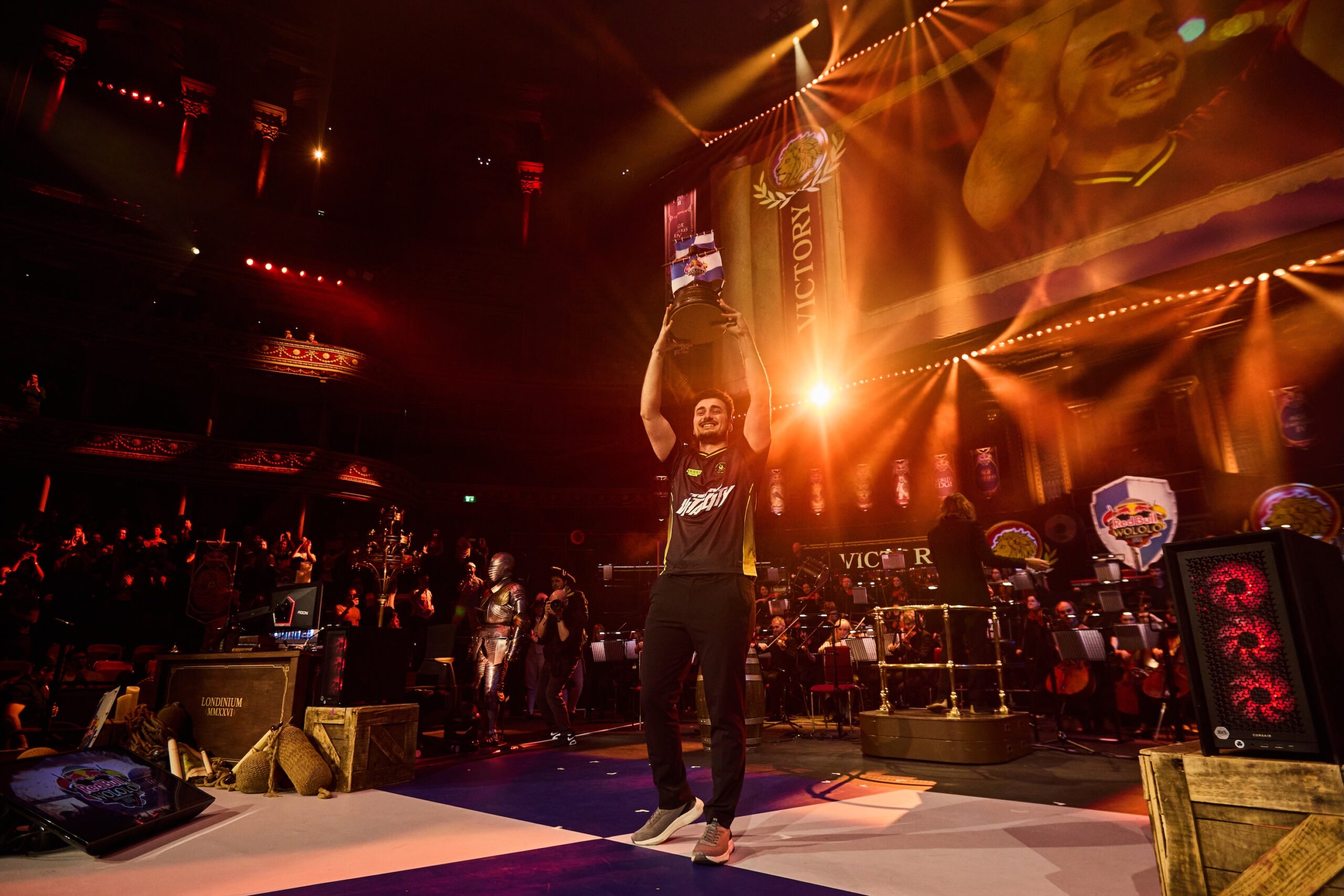 A person in esports attire stands center stage holding a trophy overhead, smiling. The stage, lit with colorful lights and a Red Bull LOIO victory banner projected above, highlights their image as an enthusiastic audience watches in the background.