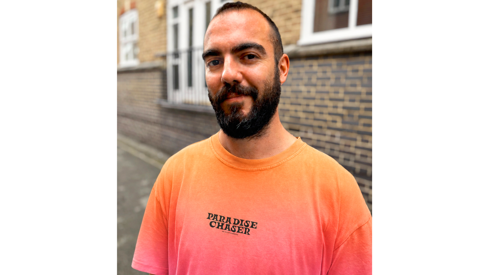 A man with a beard and short hair stands outside by a brick wall, wearing an orange gradient T-shirt featuring a creative design with the text PARADISE CHASER printed on it.
