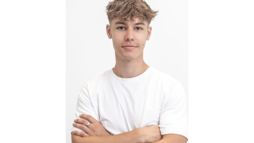 Young person with short, tousled hair stands against a plain white background, wearing a white t-shirt that subtly hints at creative design flair, arms crossed and a neutral facial expression.