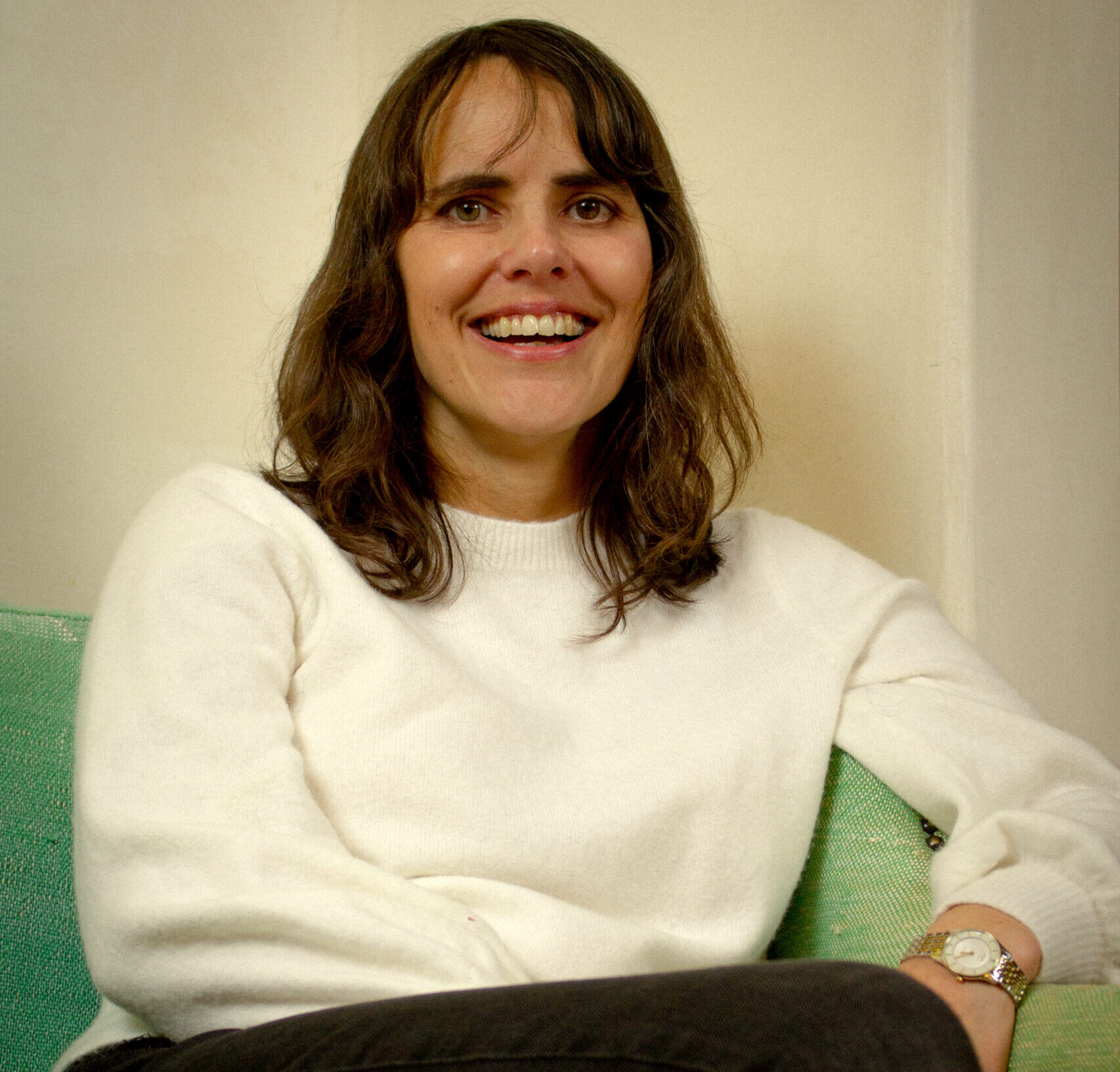 A woman with long brown hair, wearing a white sweater and a watch, sits on a green couch and smiles warmly at the camera, embodying the spirit of invention and reinvention.