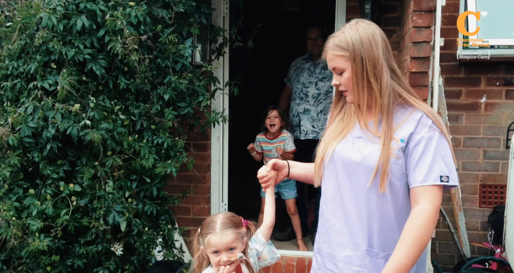 A woman in light purple scrubs holds the hand of a young girl with pigtails outside a house. Two people stand in the doorway behind them; a child smiles and an adult in glasses stands further back. Green shrubbery covers part of the scene.