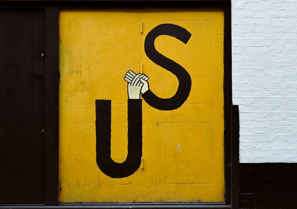 A yellow door with the letters U and S painted in black; two illustrated hands connect the tops of the letters, forming a link between them. The surrounding wall is white and dark brown.