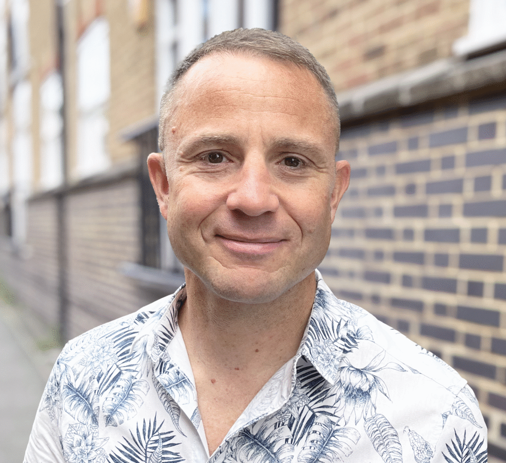 A man with short hair, wearing a white shirt with a blue leaf pattern, stands outside near a brick wall, smiling gently at the camera—a subtle storytelling strategy unfolds in the softly blurred background.