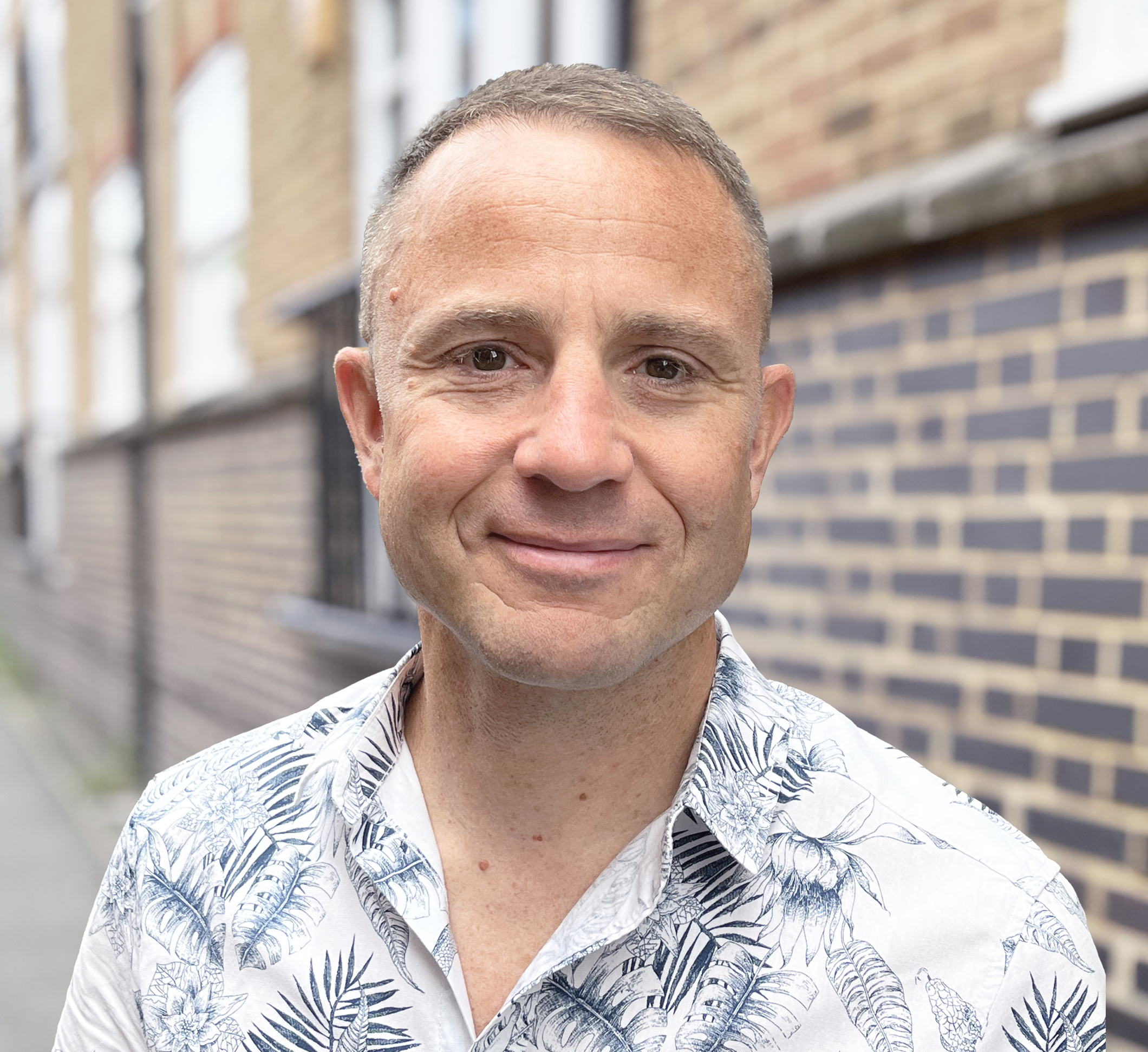 A man with short hair, wearing a white shirt with a blue leaf pattern, stands outside near a brick wall, smiling gently at the camera—a subtle storytelling strategy unfolds in the softly blurred background.
