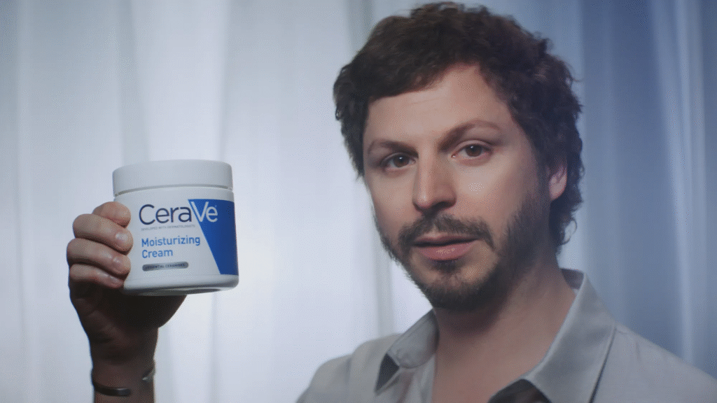 A man with short brown hair and a trimmed beard holds up a large container of CeraVe Moisturizing Cream in his right hand, standing in front of softly lit white curtains.