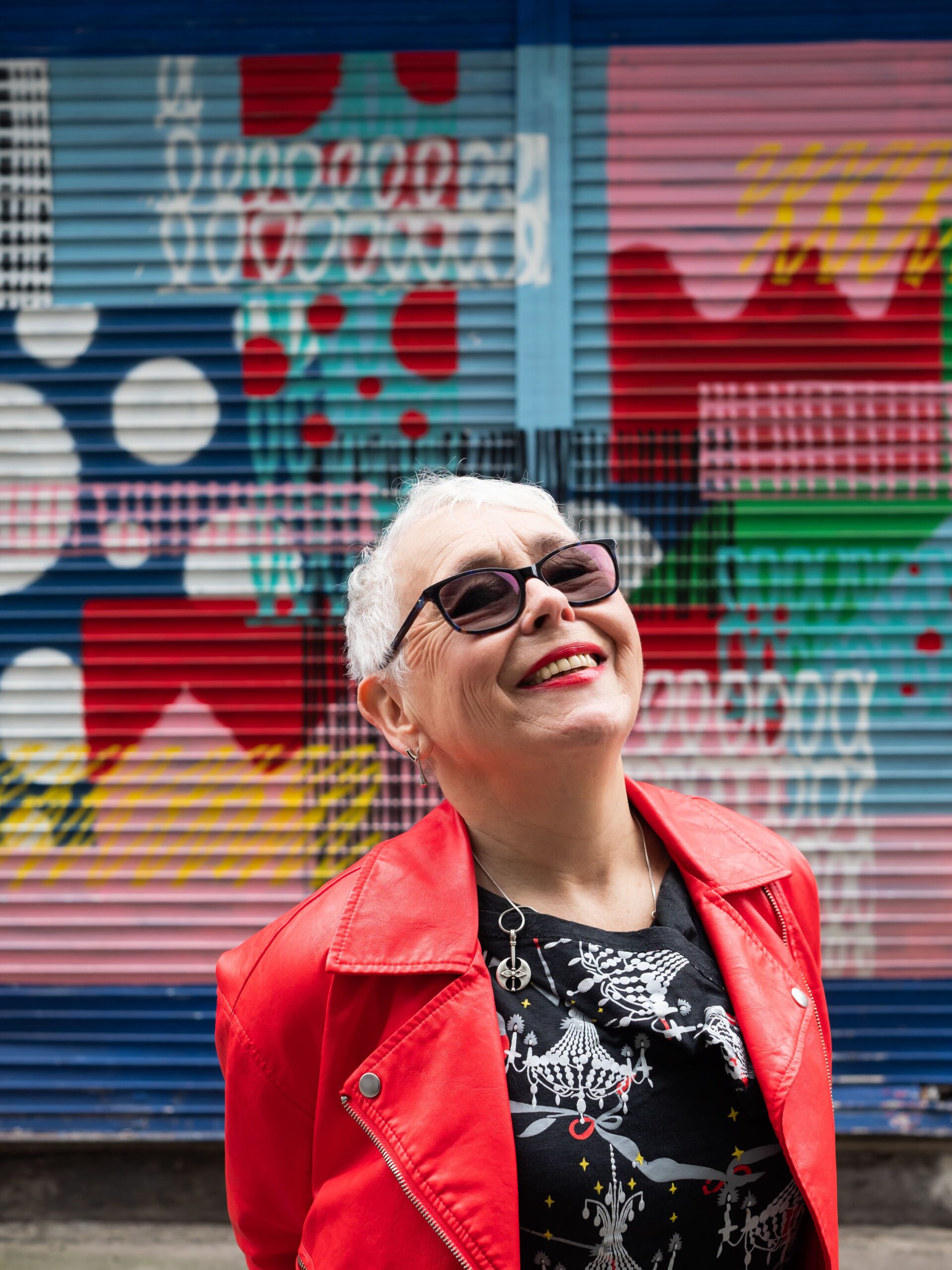 An older woman with short white hair, wearing sunglasses and a bright red jacket, smiles widely in front of a colorful, patterned mural on a metal shutter—capturing the vibrant spirit celebrated in over‑50s marketing.