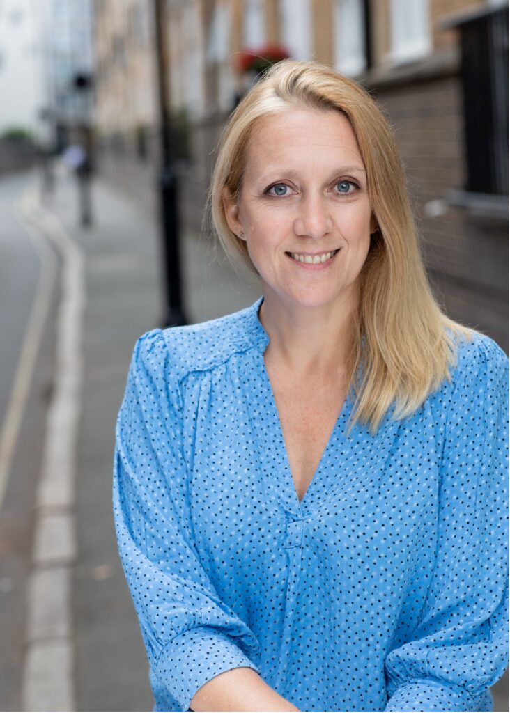 A woman with long blonde hair, wearing a light blue blouse with a dotted pattern, smiles while standing on a city sidewalk with brick buildings in the background.