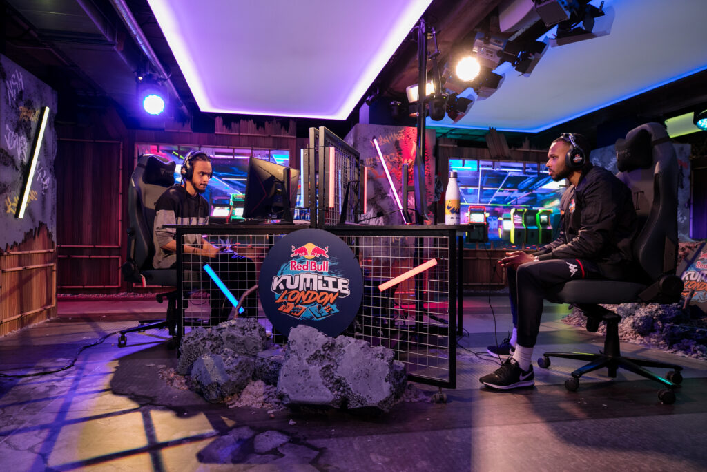 Two men sit across from each other at gaming stations in a neon-lit room, competing in an esports event with a “Red Bull Kumite London” sign on the table between them. Gaming monitors and arcade machines are visible in the background.