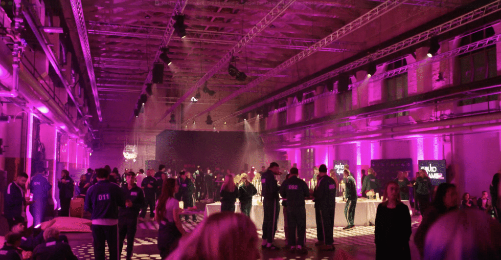 Large indoor event space filled with people socializing under vibrant pink and purple lighting, with high ceilings, visible beams, and tables scattered throughout the room—ideal for gatherings or trying out new activation tips.