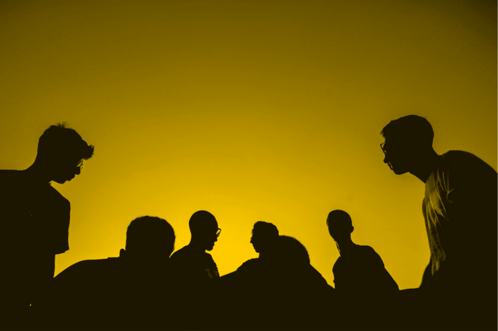 Silhouettes of six people standing and facing each other against a bright yellow, sunset-like sky, creating a dramatic and moody scene.