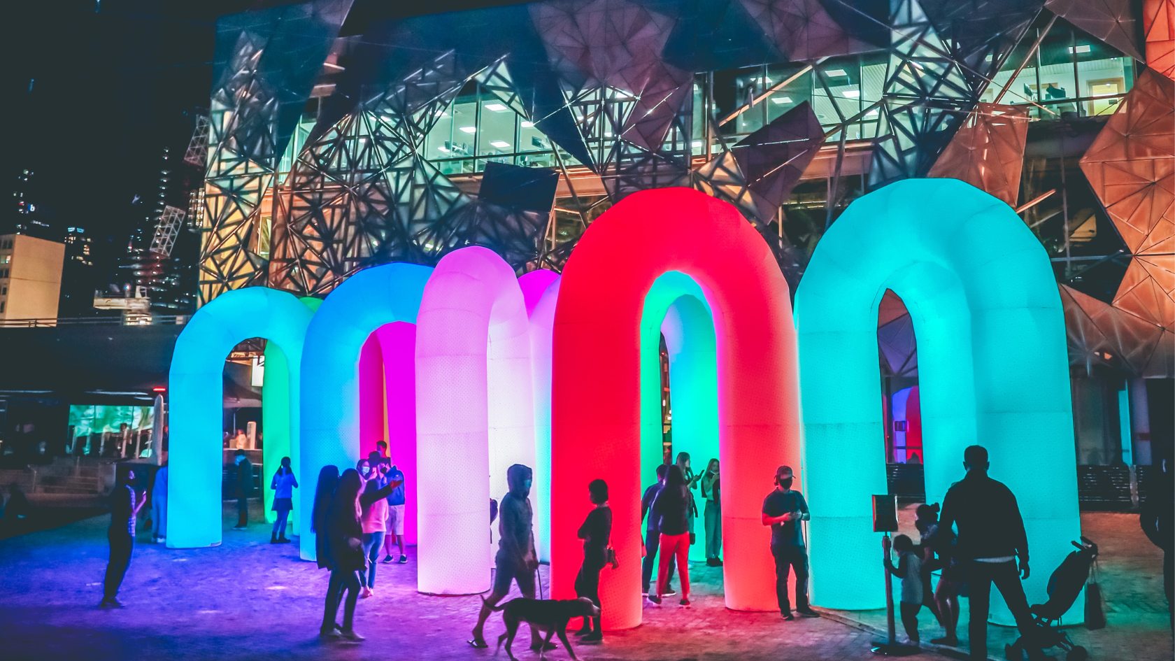 People stand and walk around large, glowing, colorful arch structures in an outdoor urban area at night, with a modern glass building in the background. The arches are lit in blue, pink, and red tones.