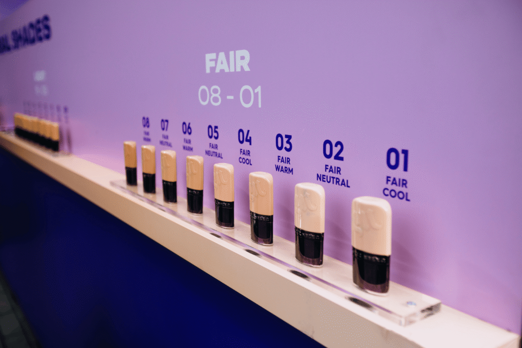 Ten foundation bottles are displayed in a row against a purple background, each labeled with a shade number and tone description, from 01 FAIR COOL to 08 FAIR COOL, ready for your perfect match at our upcoming live activation.