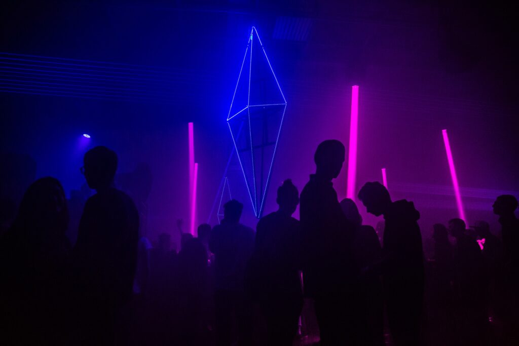 Silhouettes of people gather at a dark nightclub with bright pink and blue neon lights and a geometric, illuminated structure hanging above the crowd.