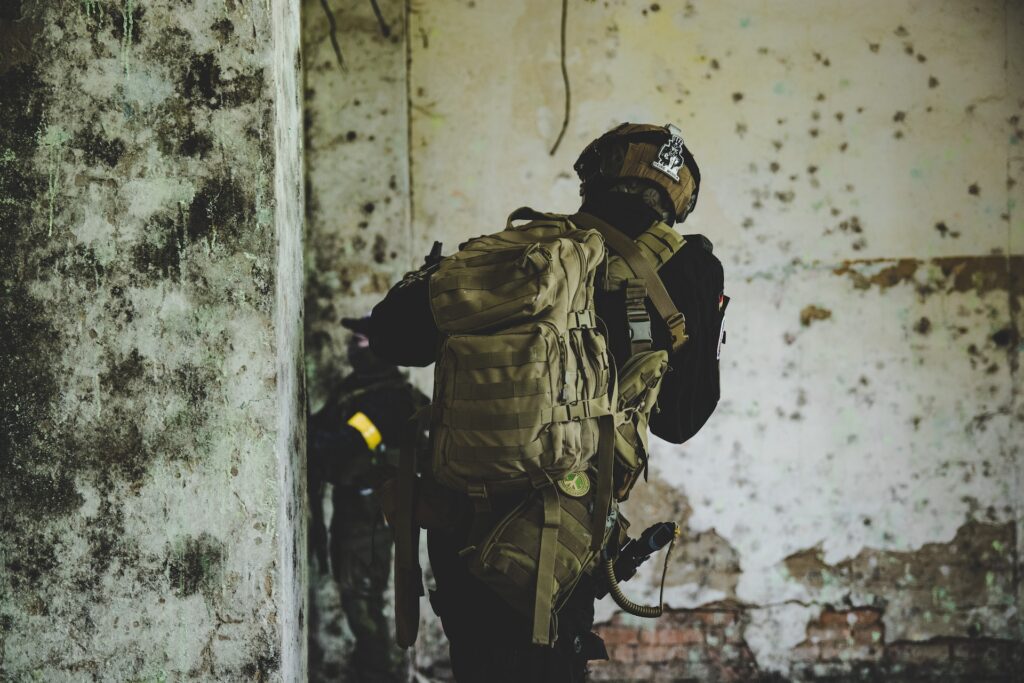 A soldier in tactical gear and a helmet, carrying a large backpack, moves cautiously through a dilapidated, moldy building—an environment ideal for experiential simulations. Another soldier is partially visible in the background.